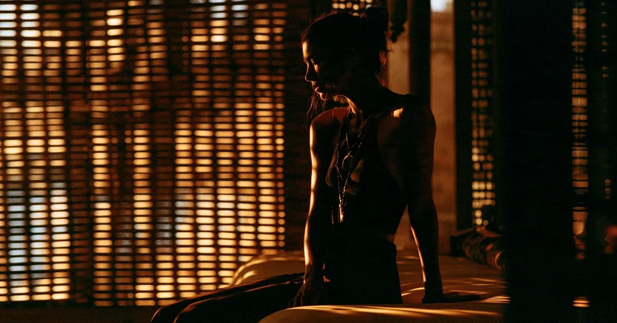 A woman leaning on the edge of a low bed in a dim room, one hand resting down to steady herself, warm golden light through slatted blinds behind her